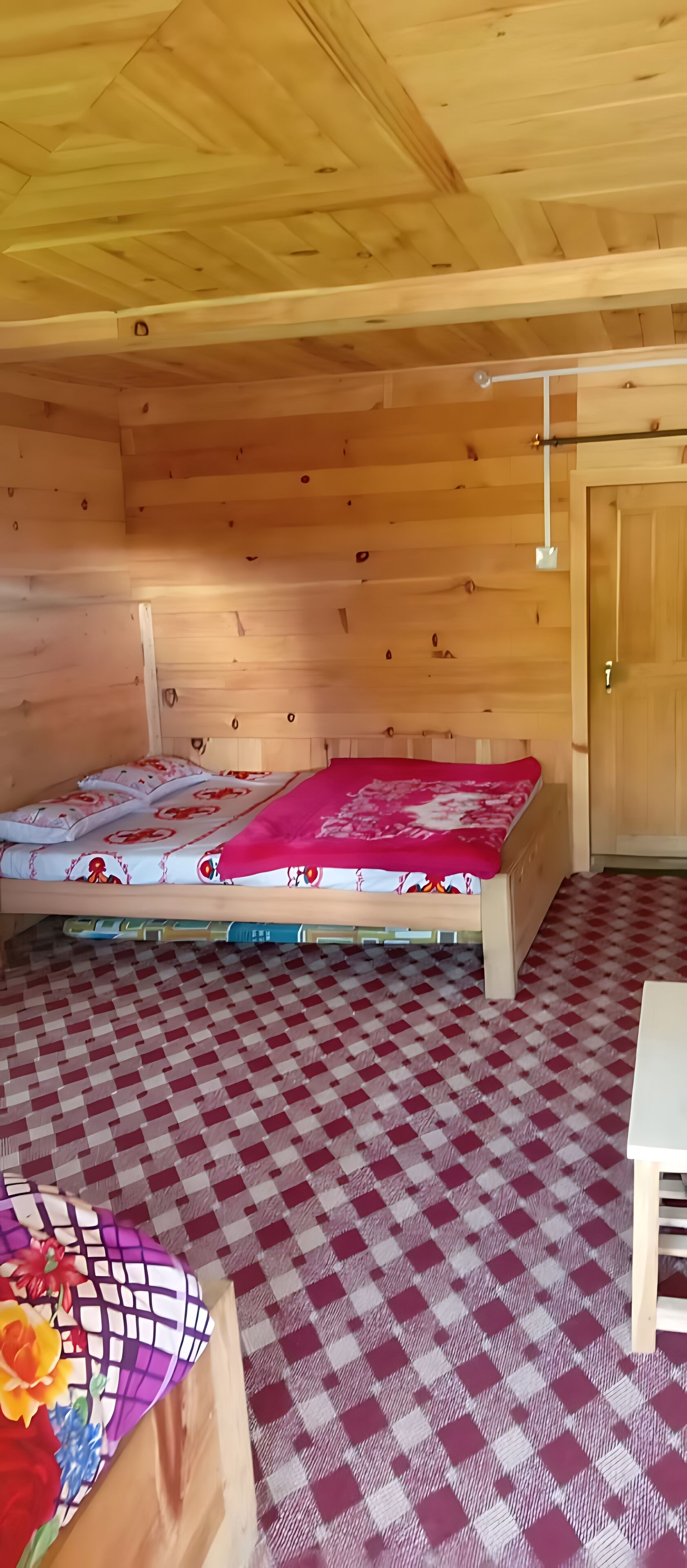 A wooden cabin room with a bed, a checkered floor, and a table with a vase of flowers.
