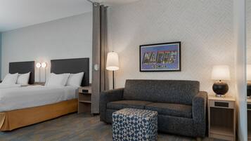 Una habitación de hotel con una cama, un sofá, un reposapiés estampado, un cuadro colgado en la pared y una mesita de noche con una lámpara.