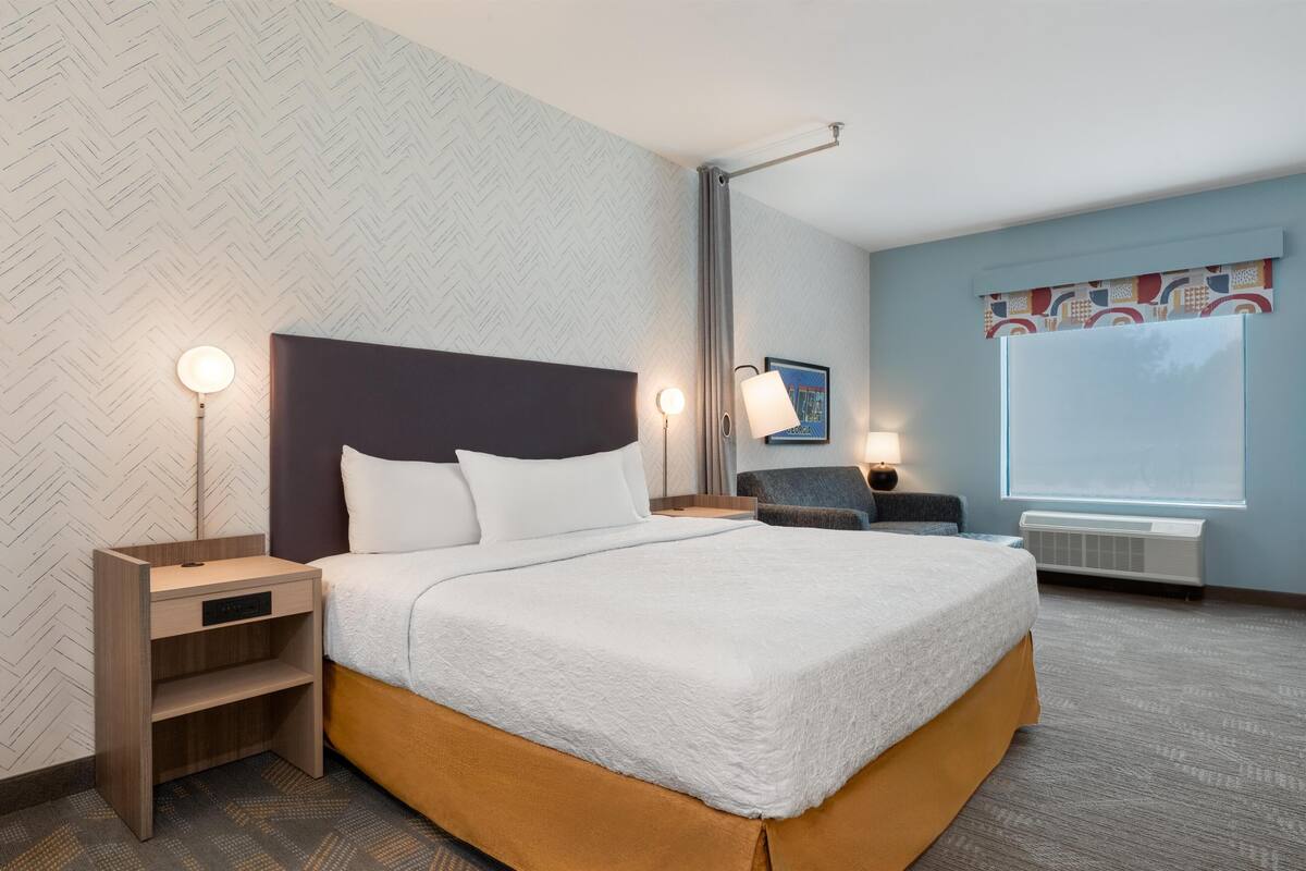 Une chambre d’hôtel comprenant un lit, des tables de chevet, une lampe, une chaise, une fenêtre et un mur orné d’un motif en chevrons.