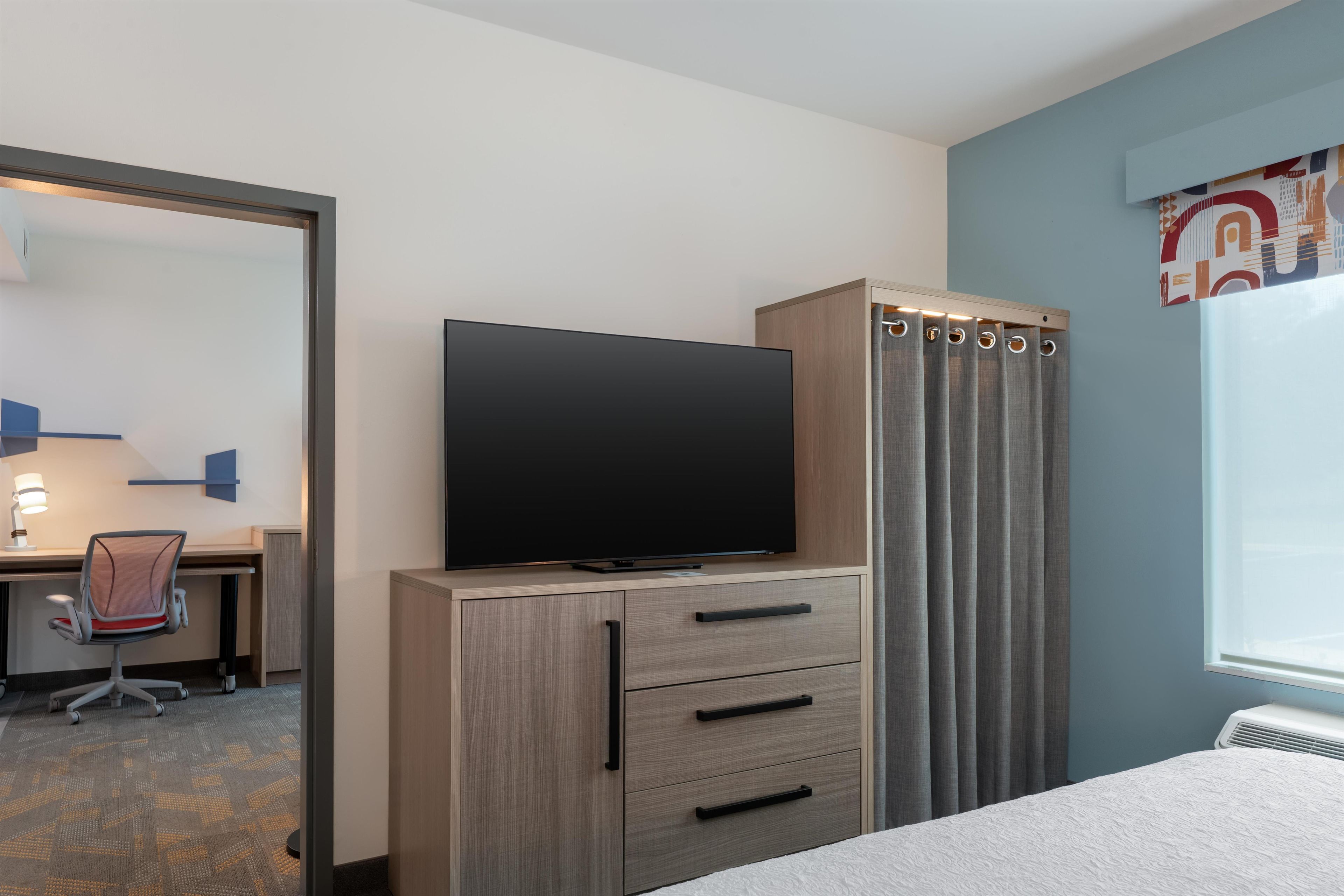 Ein Hotelzimmer mit einem Flachbildfernseher auf einer Holzkommode, einem Schreibtisch mit Stuhl sowie einem Bett mit weißer Strukturdecke.