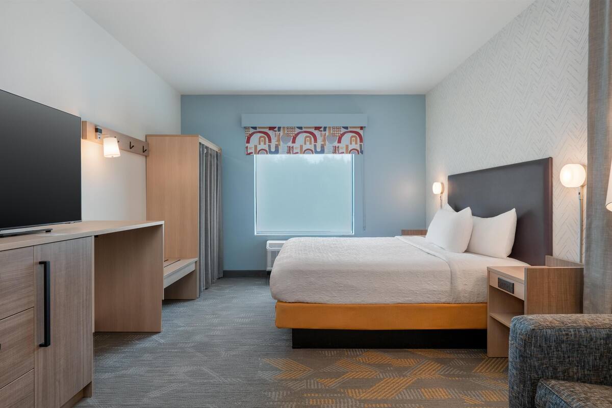 Une chambre d’hôtel de taille réduite, comprenant un lit, des tables de chevet, un téléviseur, une armoire et une fenêtre agrémentée d’un rideau à motifs.