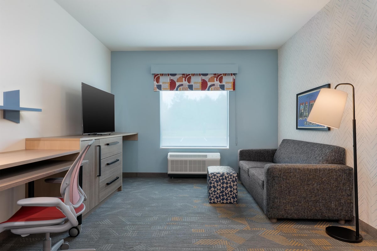 Une chambre d’hôtel de taille modeste, équipée d’un bureau, d’une chaise, d’une télévision, d’un canapé et d’un tapis à motifs.