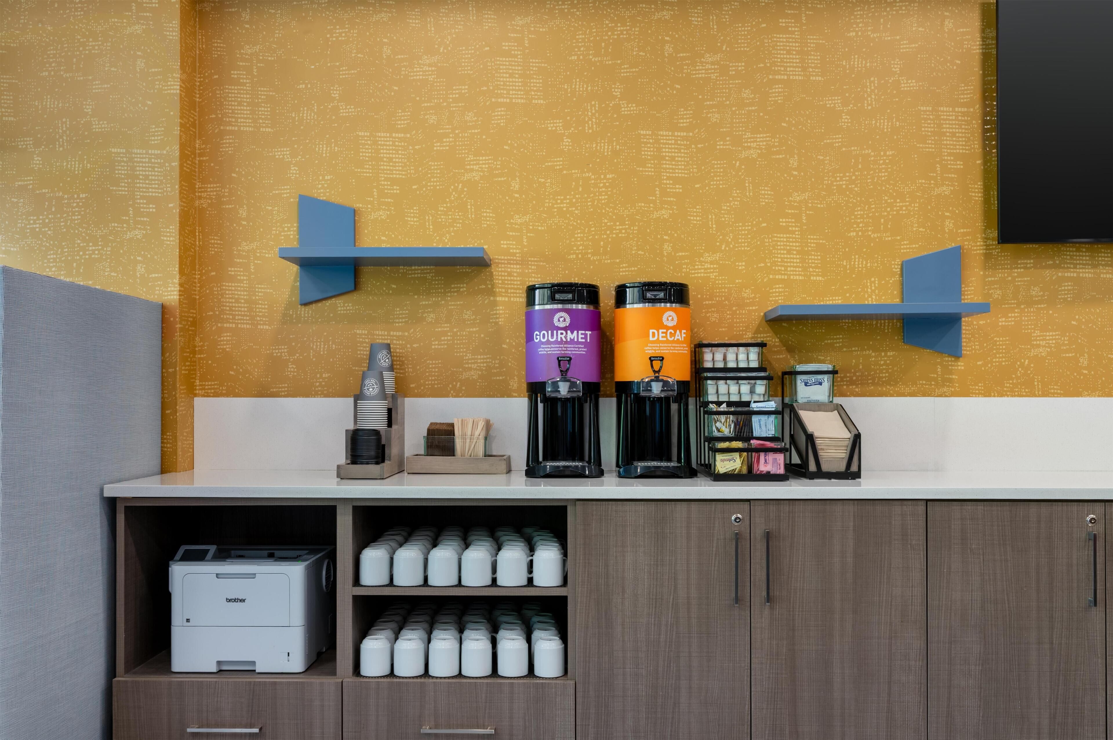 Un coin café équipé de deux machines à café, l’une portant la mention « GOURMET » et l’autre « DÉCAFÉINÉ », d’une machine à café et d’une imprimante, le tout installé sur un comptoir surmonté d’un meuble en bois.