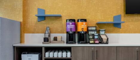 A coffee station with two coffee makers labeled 'GOURMET' and 'DECAF,' a coffee machine, and a printer on a counter with a wooden cabinet below.