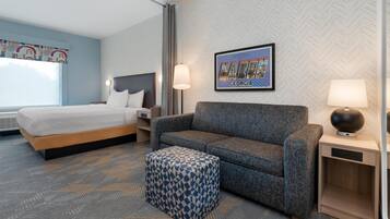 Habitación de hotel con cama, sofá, reposapiés estampado, mesita de noche con lámpara, y pared con diseño de espiga y un cuadro enmarcado.