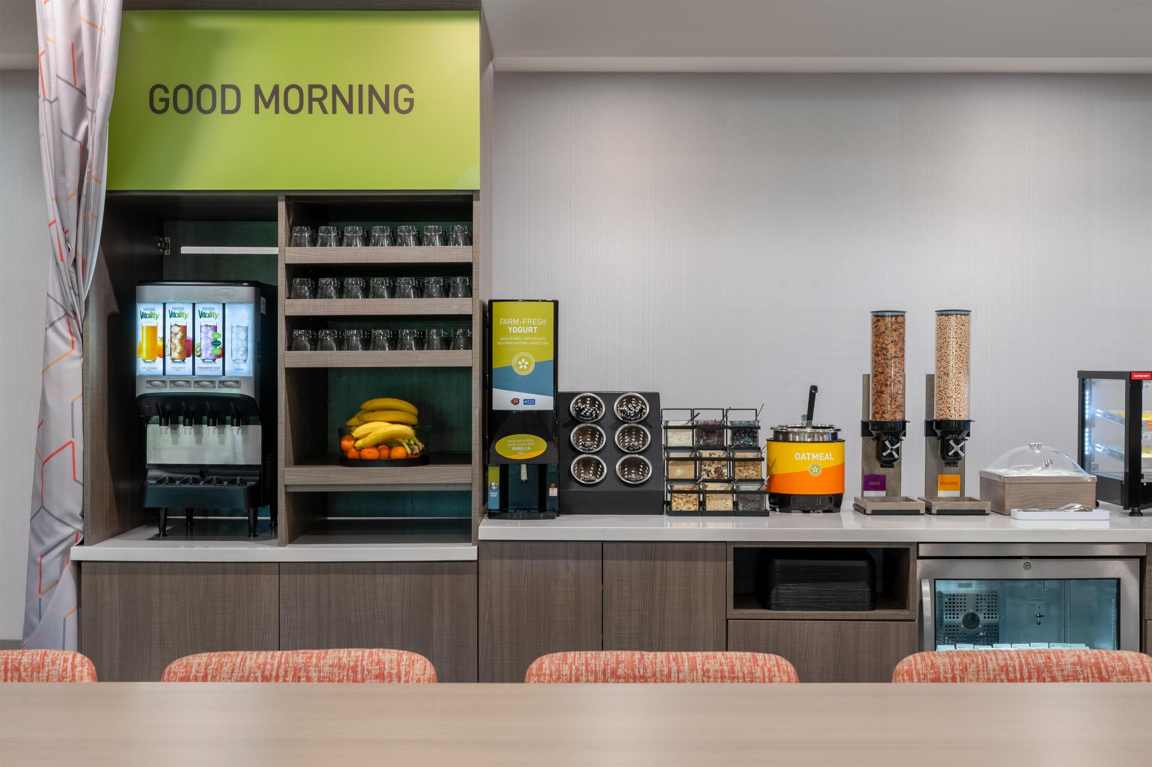 Una barra de desayuno con un letrero que dice 'GOOD MORNING', una máquina de café, una exhibición de frutas y varios recipientes de comida.