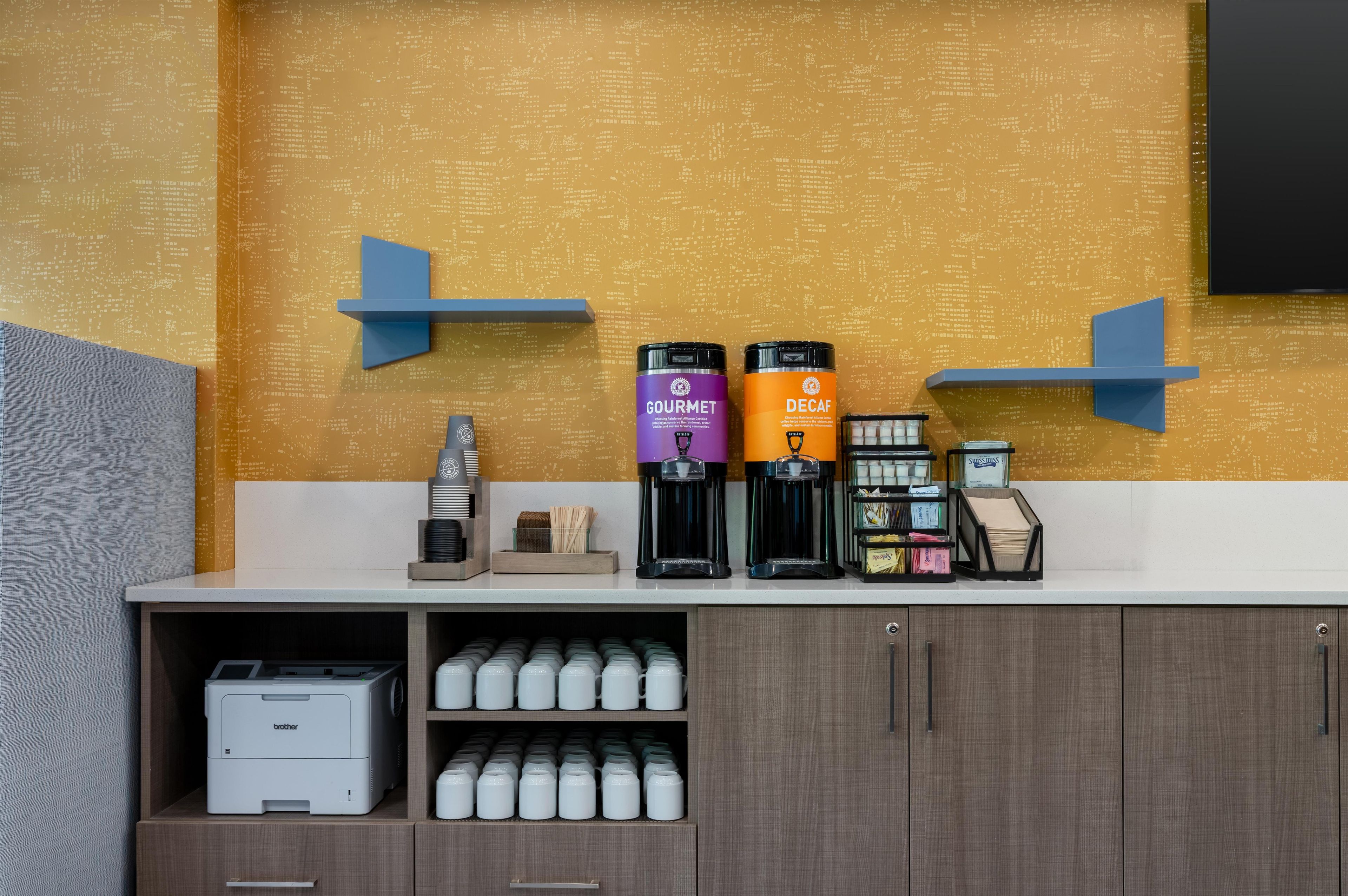 A coffee station with two coffee machines labeled 'GOURMET' and 'DECAF' and a coffee maker. There are stacks of disposable cups and a printer on the counter.