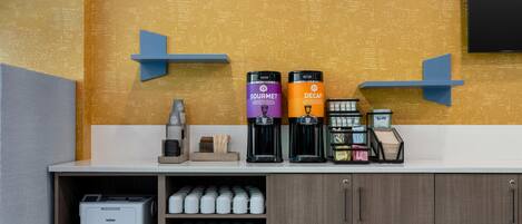 A coffee station with two coffee machines labeled 'GOURMET' and 'DECAF' and a coffee maker. There are stacks of disposable cups and a printer on the counter.