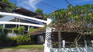 A two-story building with a white facade, a balcony, and a tree with pink flowers in the foreground. - Villa Joglo Paris Yogyakarta (Parangtritis)