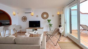 A modern living room with a flat-screen TV, a sofa, a dining area with chairs, and a balcony with a chair.