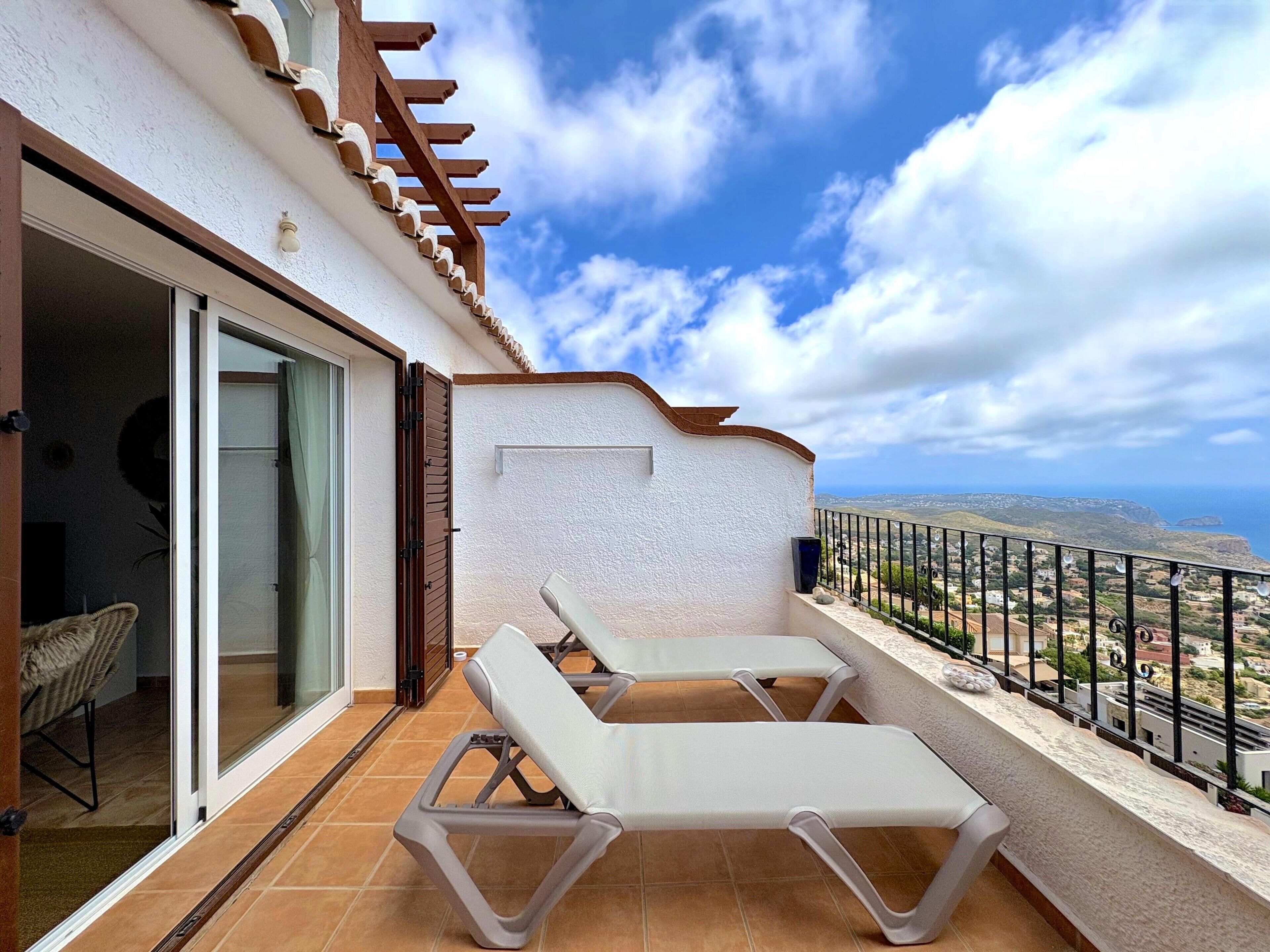A balcony with two white lounge chairs, a glass door leading to an interior space, and a view of the coastline.