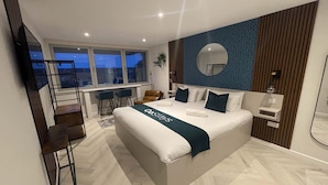 A modern bedroom with a large bed, a view of the city, a desk with chairs, and a small side table.