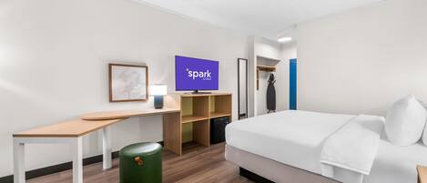 A modern hotel room with a bed, desk, chair, TV, and storage shelves.