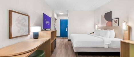 Quarto de hotel com cama, escrivaninha, cadeira, abajur, lixeira e televisão na parede.
