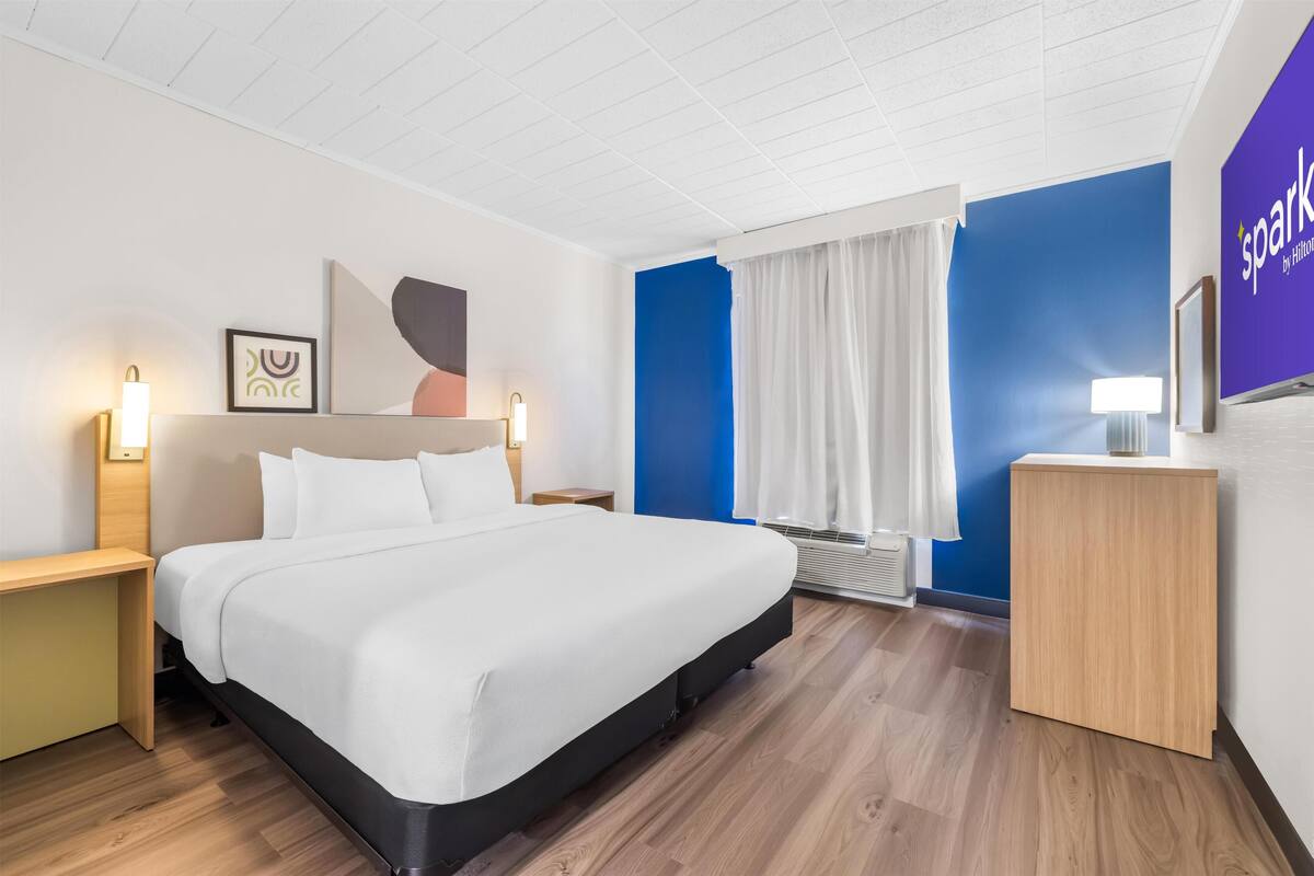 Une chambre d’hôtel comprenant un lit, des tables de chevet, un bureau, un téléviseur fixé au mur et une fenêtre avec des rideaux.