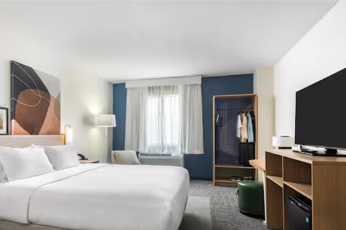 Una habitación de hotel con cama, mesitas de noche, televisor, armario y ventana con cortinas.