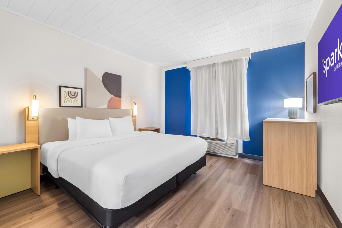 Habitación de hotel con cama, mesitas de noche, escritorio, televisor montado en la pared y ventana con cortinas.