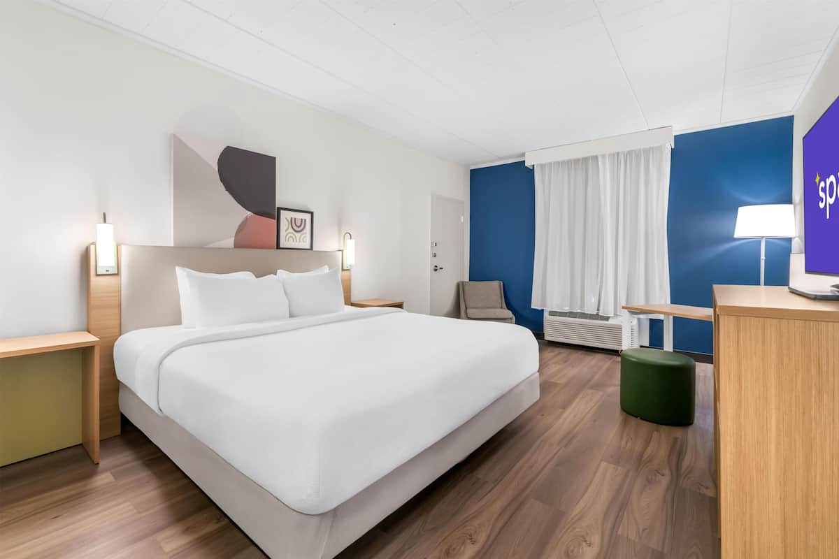 Une chambre d’hôtel comprenant un lit, des tables de chevet, un bureau, une chaise, un pouf vert, un climatiseur fixé au mur et une fenêtre avec des rideaux.