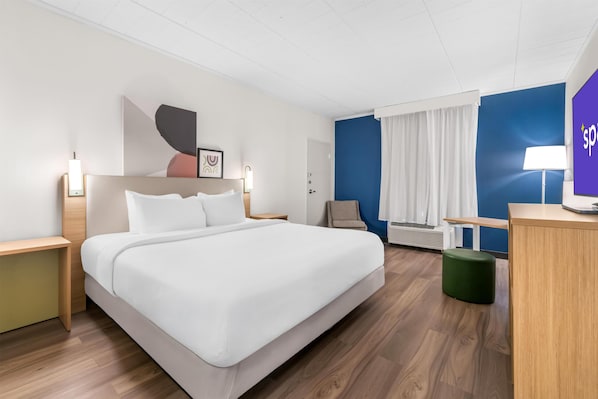 A hotel room with a bed, bedside tables, a desk, a chair, a green ottoman, a wall-mounted air conditioner, and a window with curtains.