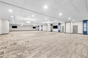 A spacious room with a large carpeted floor, white walls, and a grid of ceiling lights.