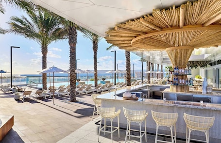 A rooftop bar with a view of the ocean, palm trees, and a pool.