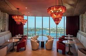 Un salón moderno con mesas redondas y sillas rojas, amplias ventanas que ofrecen vistas a la ciudad y lámparas decorativas colgantes.
