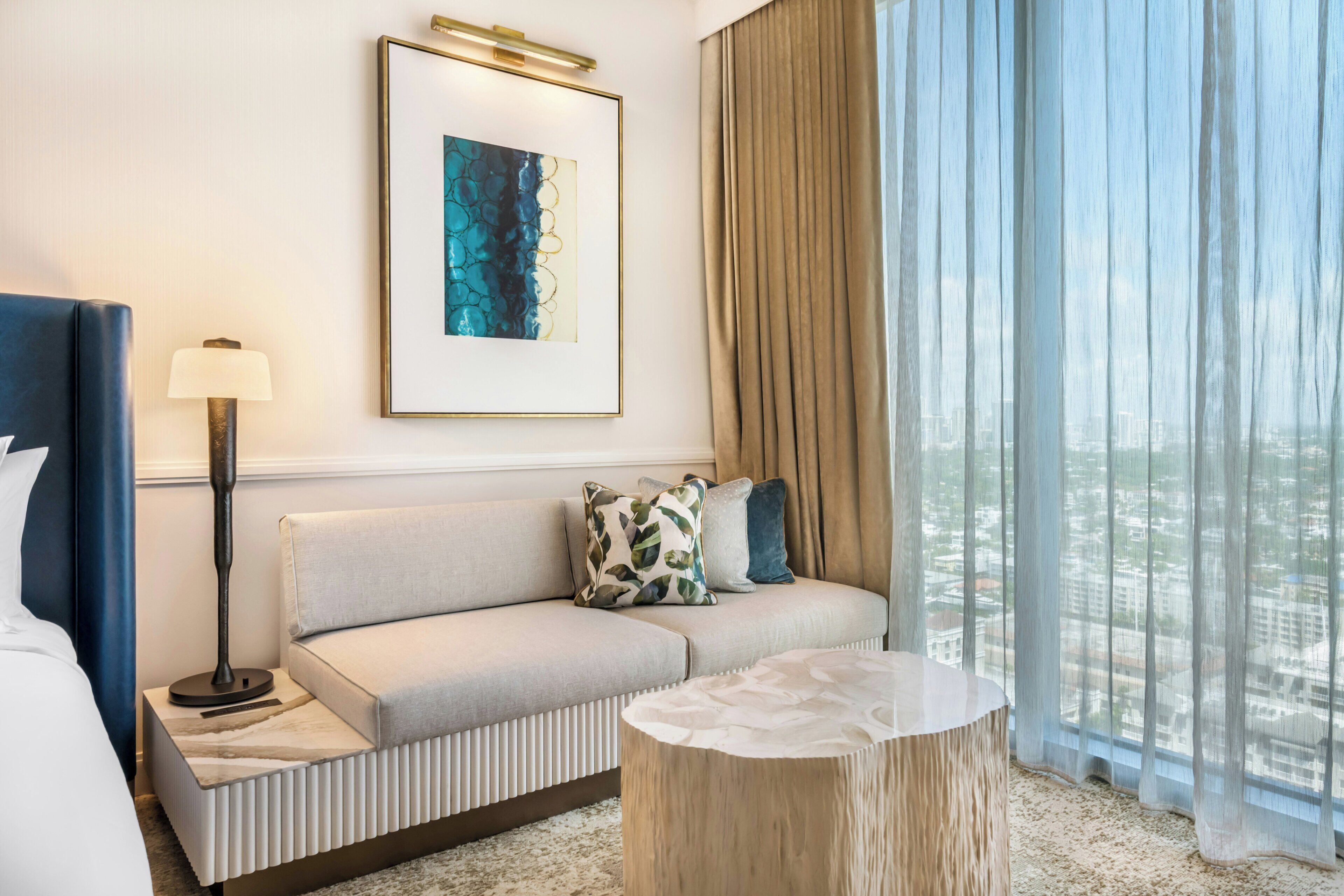 A modern hotel room with a sofa, a round wooden side table, a framed abstract artwork, and a large window with sheer curtains.