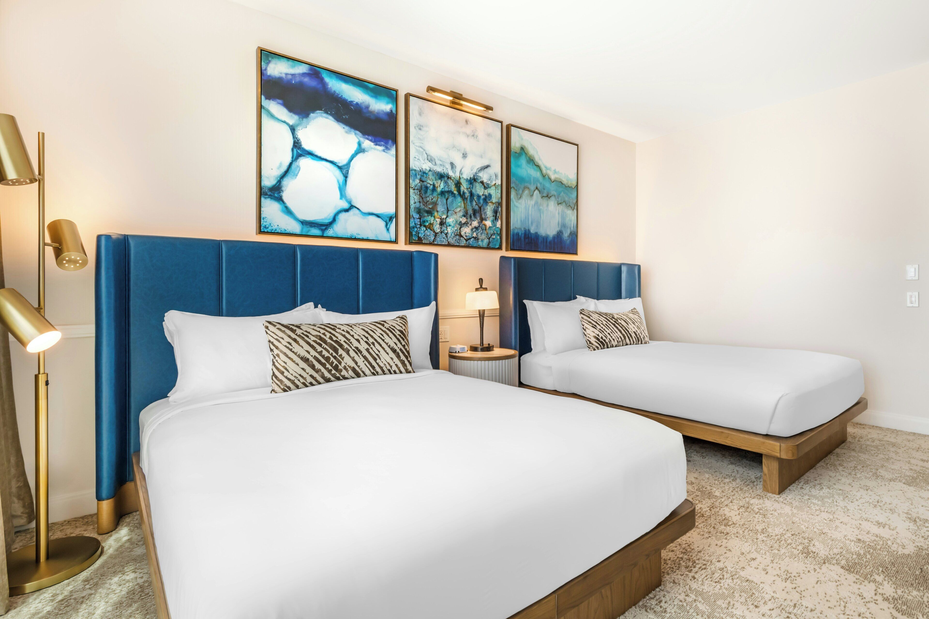 A hotel room with two beds, a nightstand, a lamp, and abstract wall art.