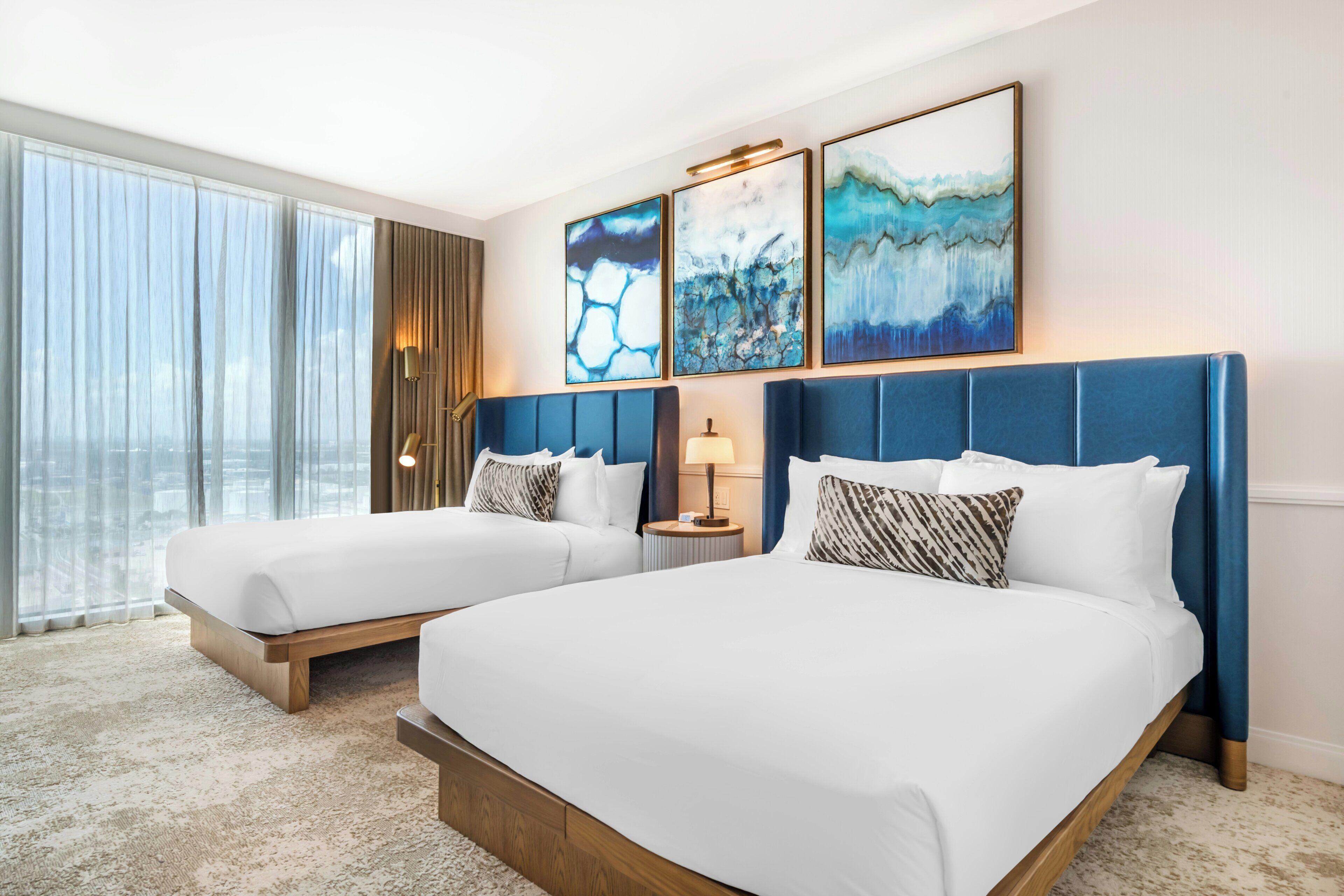 A hotel room with two beds, a large window with curtains, and three abstract paintings on the wall.