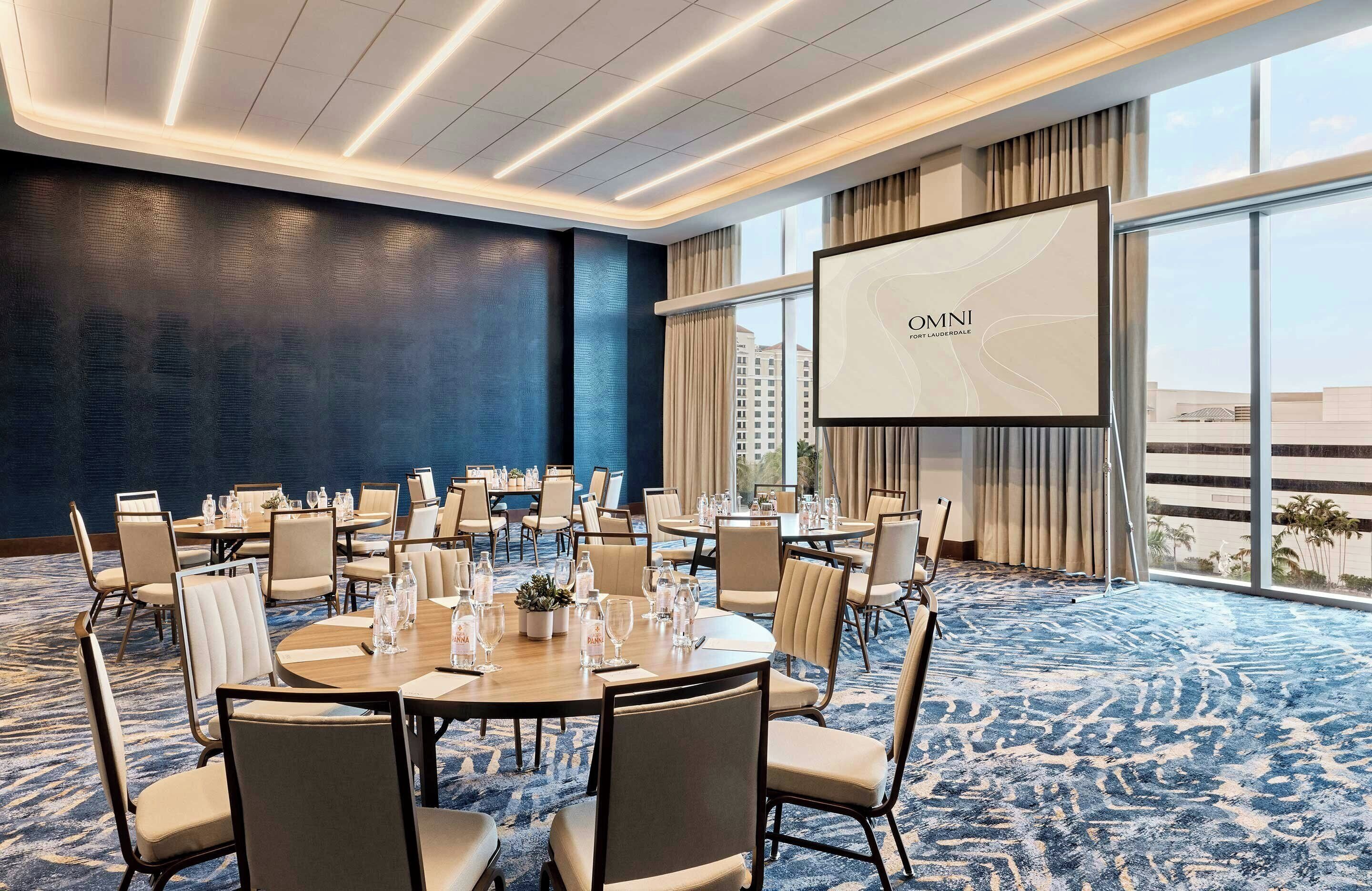 A conference room with round tables, chairs, a large screen, and a view of buildings through large windows.