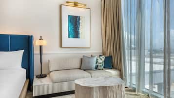 A hotel room with a bed, a sofa, a side table, a framed artwork, and a large window with curtains.