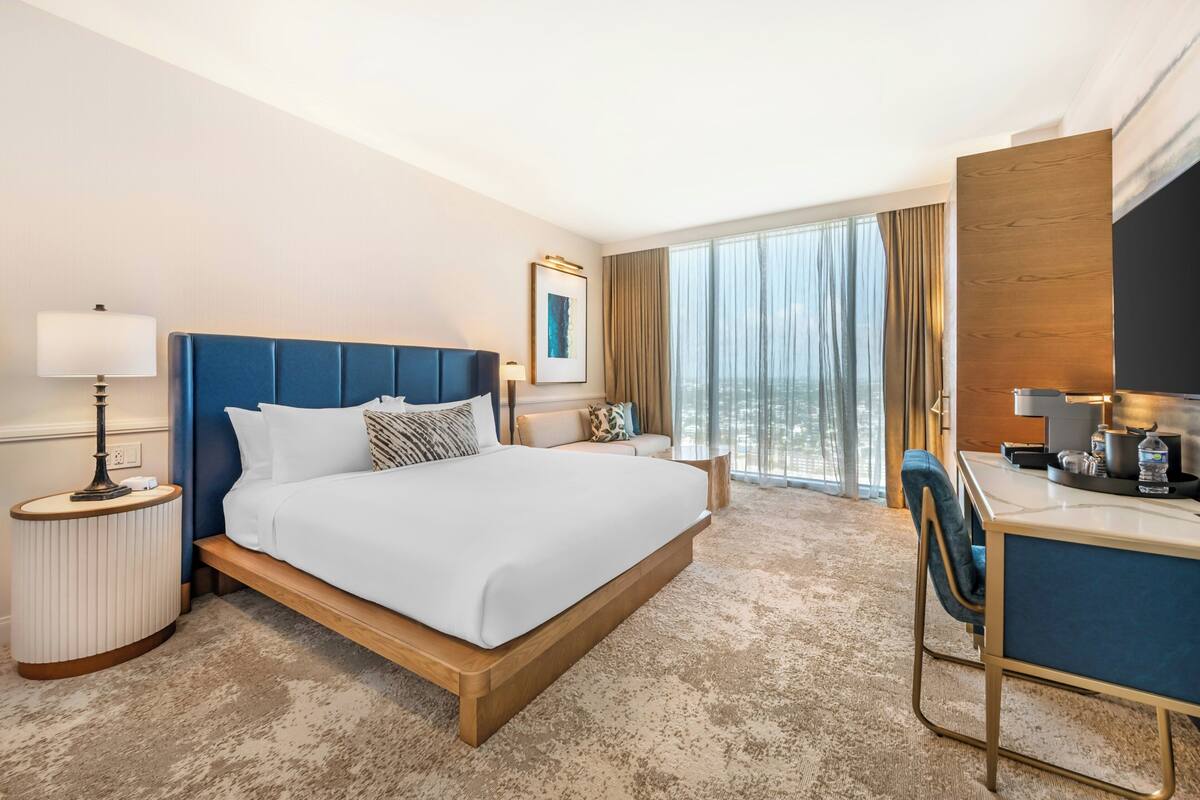 Une chambre d’hôtel comprenant un lit, une table de chevet, une lampe, un bureau, une chaise et une grande fenêtre avec des rideaux.