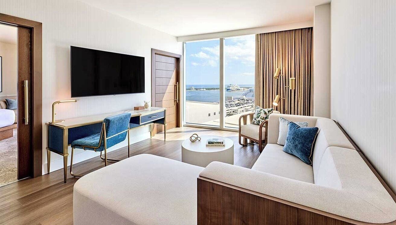 A modern hotel room with a large flat-screen TV, a sofa, a desk with a chair, and a large window offering a view of the sea.