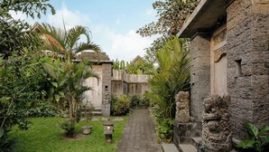 A stone pathway leading through a lush garden with tropical plants and a stone building with intricate carvings. - Kumala Ubud Villa (Ubud)