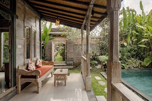 Un área de estar al aire libre con techo, una banca y una mesa de madera, un sendero de piedra y una piscina.