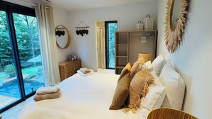 A spacious bedroom with a large bed, a wooden floor, and a view of a garden through a sliding door. - Design house in the heart of the forest Escape & absolute comfort (Roquefort-les-Pins)