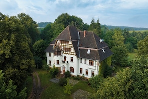 A large, white, two-story house with a steeply pitched roof, surrounded by dense trees and a well-maintained lawn. - Cozy vacation apartment for up to 4 people on the first floor of a manor house (Alt Necheln)