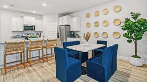 Une cuisine moderne avec des meubles blancs, un four à micro-ondes et un réfrigérateur. On y trouve un coin repas avec des chaises bleues et une table. Les murs sont décorés d’assiettes en bois de forme circulaire.