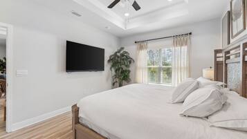 A bedroom with a bed, a ceiling fan, a TV mounted on the wall, a window with curtains, and a plant.