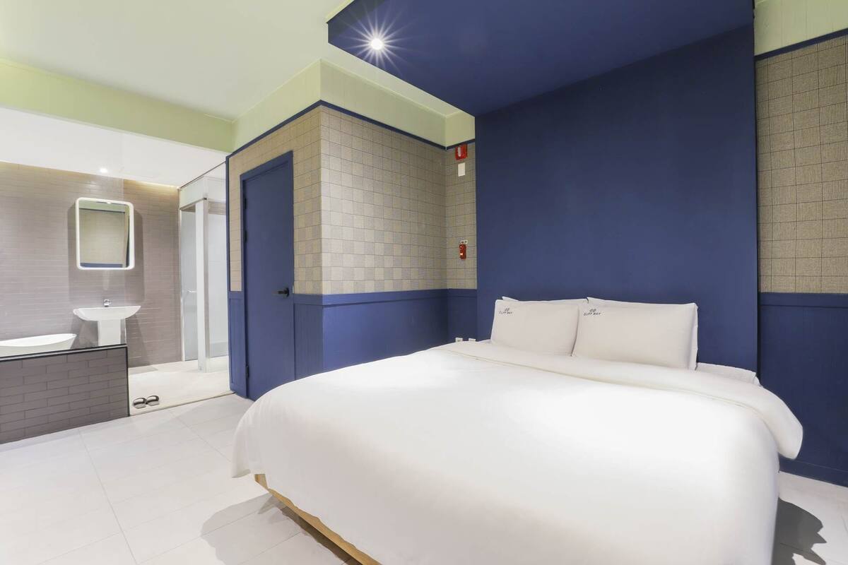 Habitación de hotel con una cama grande, un baño con lavamanos y espejo, y una pared azul de acento.
