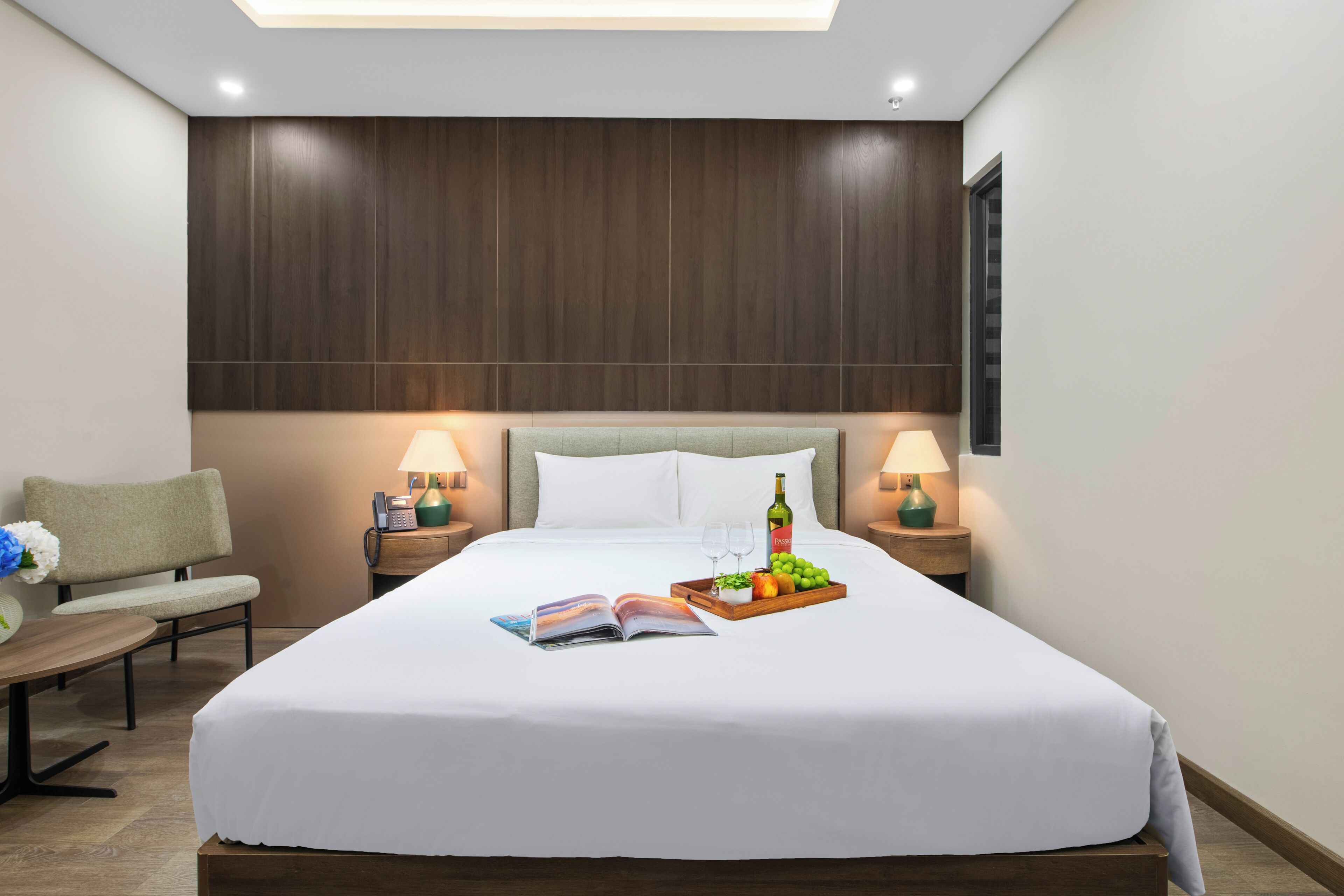 Una habitación de hotel impecable con una cama grande, lámparas de noche, una mesita de noche con un libro y una bandeja de frutas, y una silla.