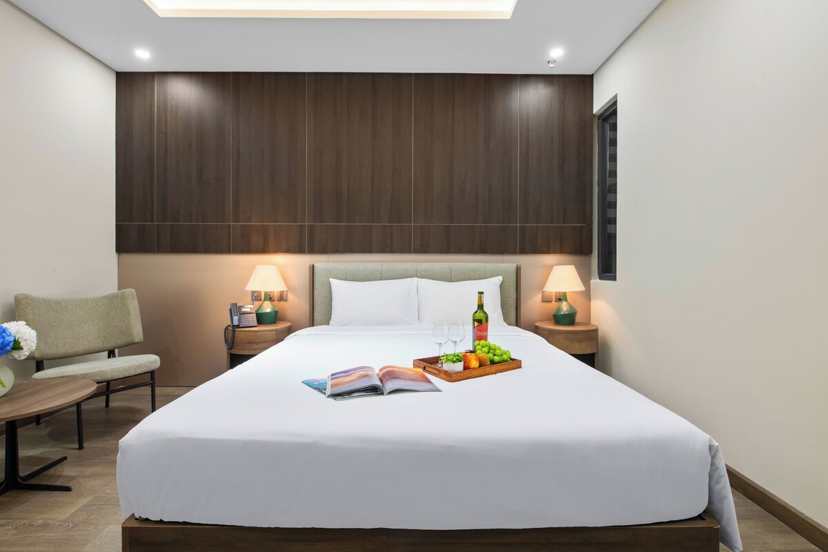 Ein ordentlich eingerichtetes Hotelzimmer mit einem großen Bett, Nachttischlampen, einem Nachttisch mit einem Buch und einer Obstplatte sowie einem Sessel.