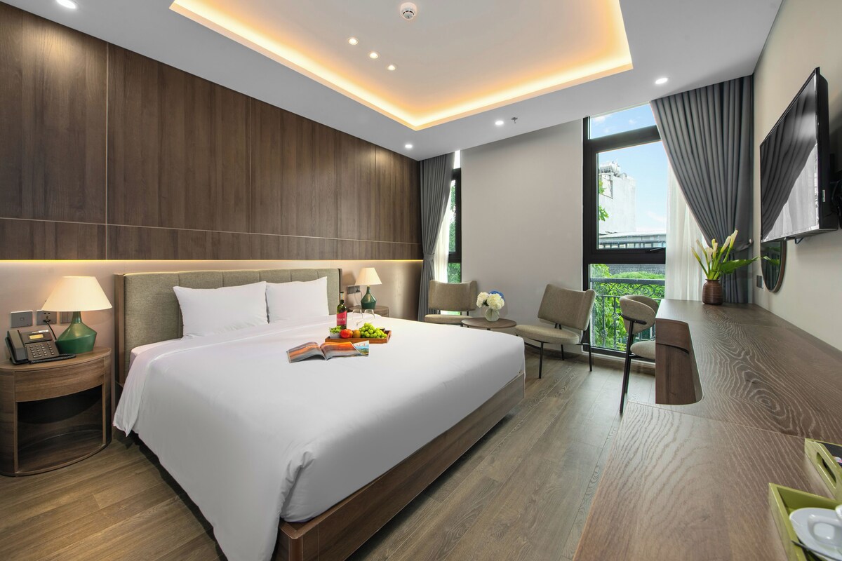 A modern hotel room with a large bed, bedside tables, a desk, and a chair. There is a TV mounted on the wall and a window with curtains.