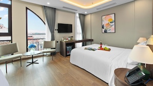 Une chambre d’hôtel avec un grand lit, un bureau, une chaise, une petite table ornée d’un vase de fleurs, un téléviseur fixé au mur et un balcon avec vue.
