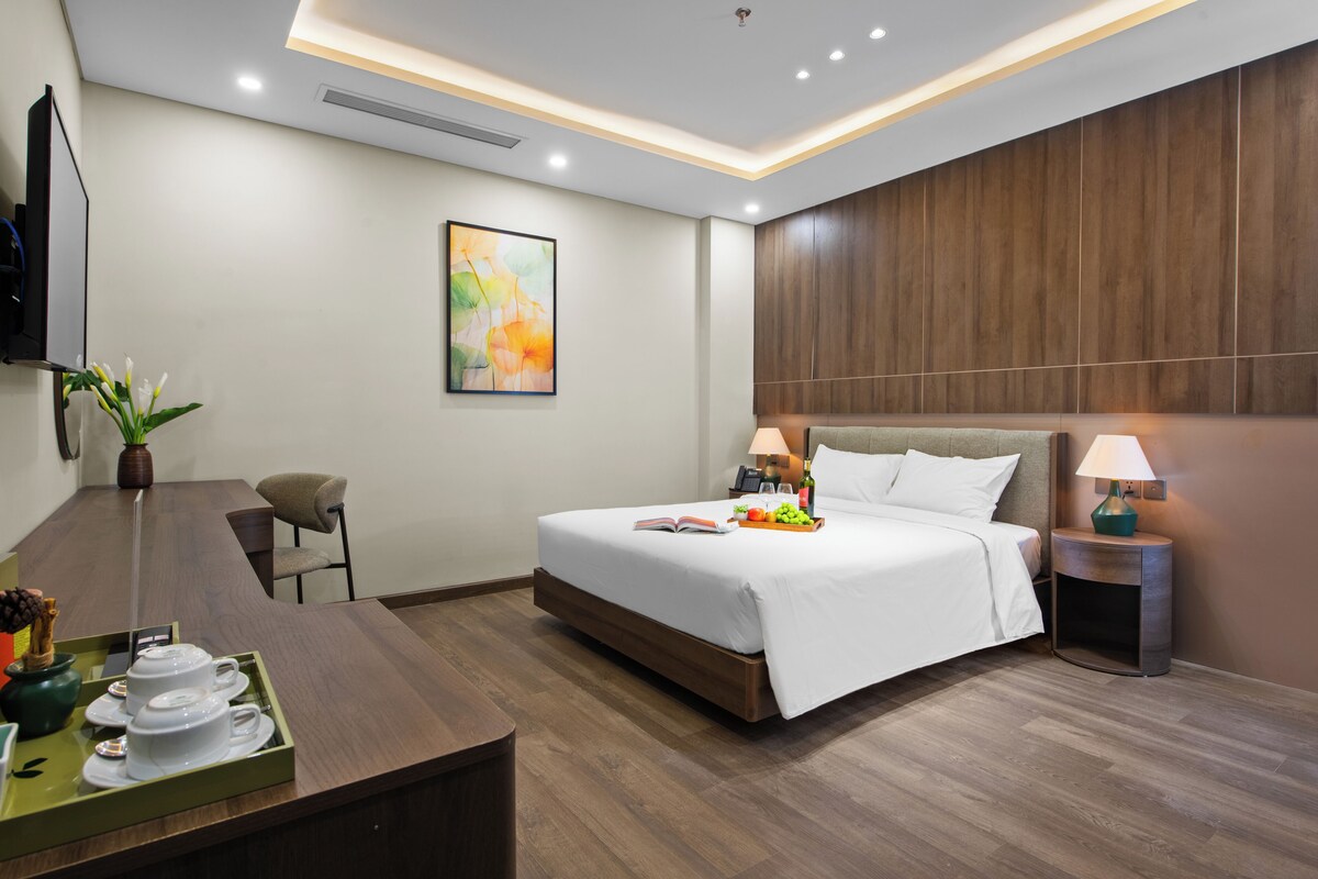 Ein modernes Hotelzimmer mit einem großen Bett, Nachttischen, einem Schreibtisch mit Frühstücksgebäck und -speisen, einem Sessel und einem an der Wand befestigten Fernseher.