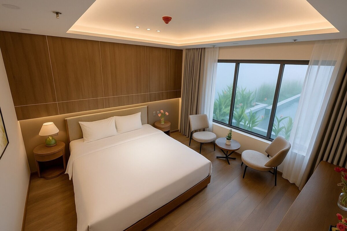 A modern hotel room with a large bed, a sitting area with two chairs, a small table, and a large window offering a view of greenery.