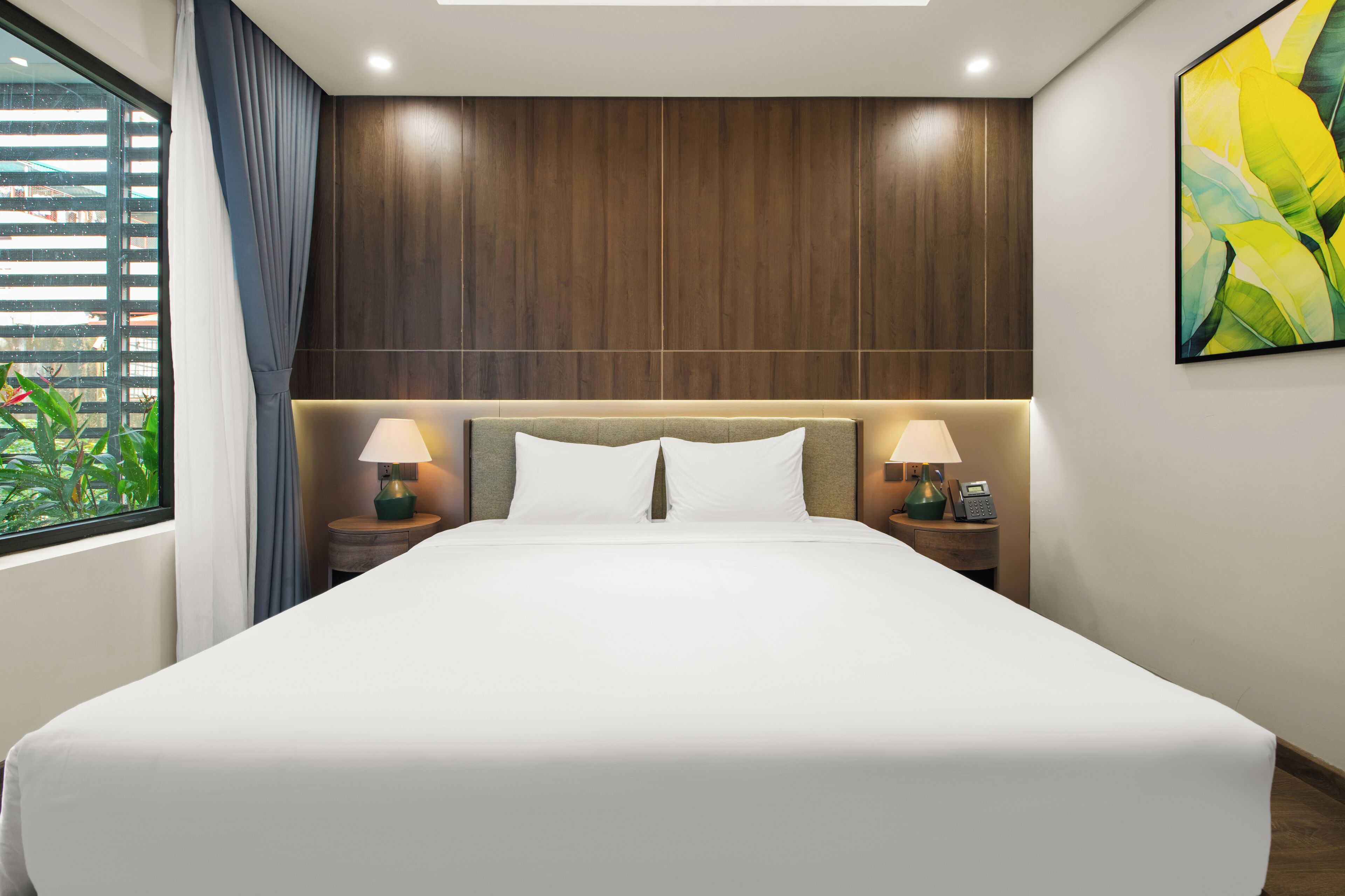 A hotel room with a large bed, wooden headboard, two bedside tables with lamps, and a window with curtains.