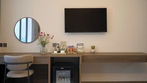 Une chambre d’hôtel moderne équipée d’un téléviseur à écran plat fixé au mur, d’un miroir rond, d’un petit réfrigérateur, d’un bureau en bois avec une chaise et d’un vase avec des fleurs.