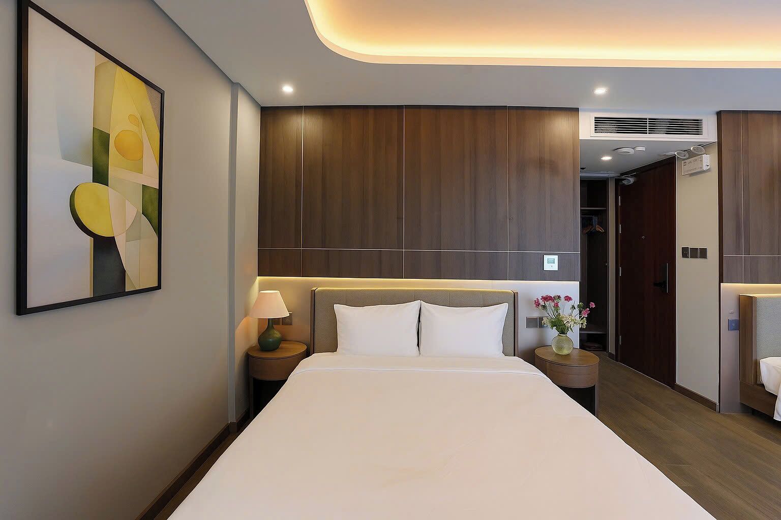 A hotel room with a large bed, wooden paneling on the walls, a bedside table with a lamp, and a framed abstract painting.