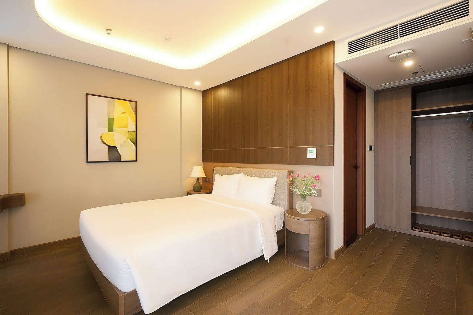 A modern hotel room with a large bed, a bedside table with a lamp, a wall-mounted artwork, and a wooden wardrobe.